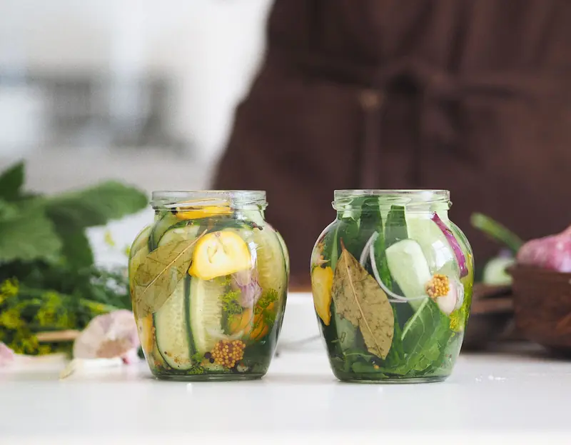 Two glass jars filled with homemade pickled cucumbers and herbs on a white kitchen surface