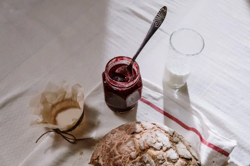 A cozy breakfast scene featuring rustic bread, a jar of jam, and a glass of milk on a textured tablecloth