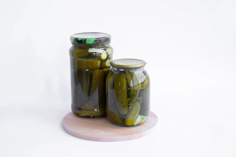 Two jars of pickled cucumbers placed on a wooden chopping board with a white background