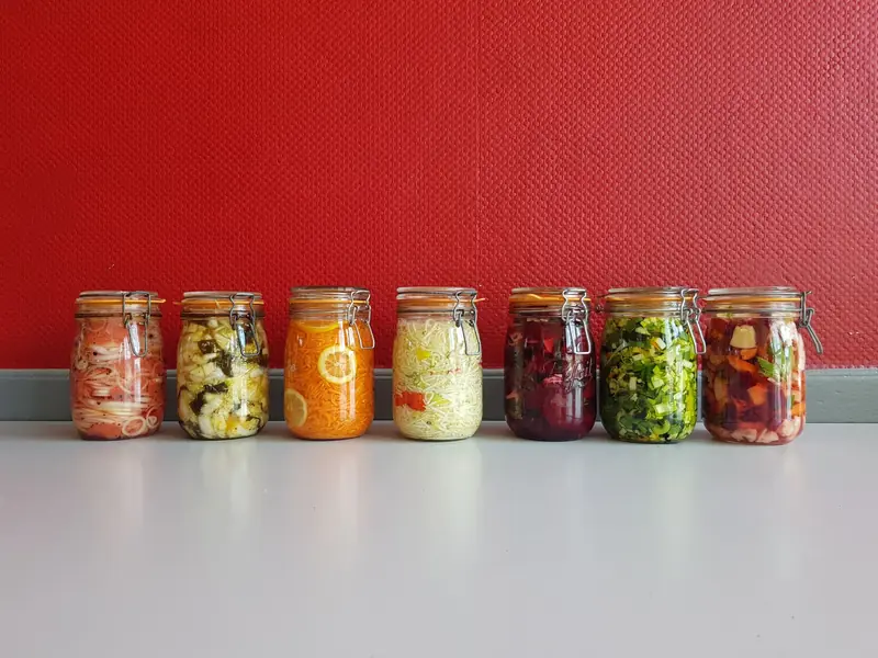 A row of vibrant, multicolored mason jars filled with assorted fermented vegetables against a red wall