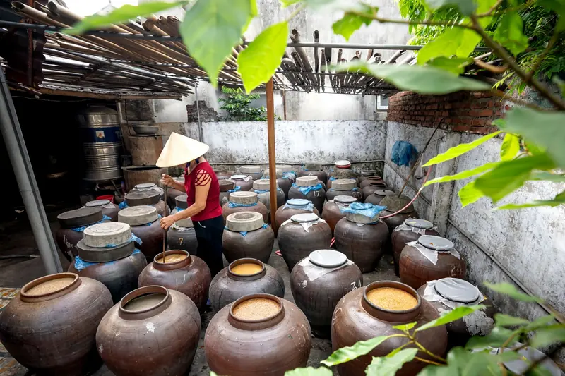 A person in a conical hat working in a traditional fermentation room with many jars