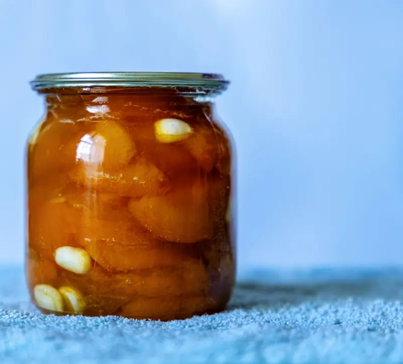 A close-up of a jar filled with preserved garlic and peaches, set against a soft blue background