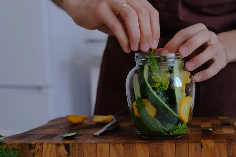 Hands organizing fresh vegetables for pickling in a glass jar on a wooden countertop